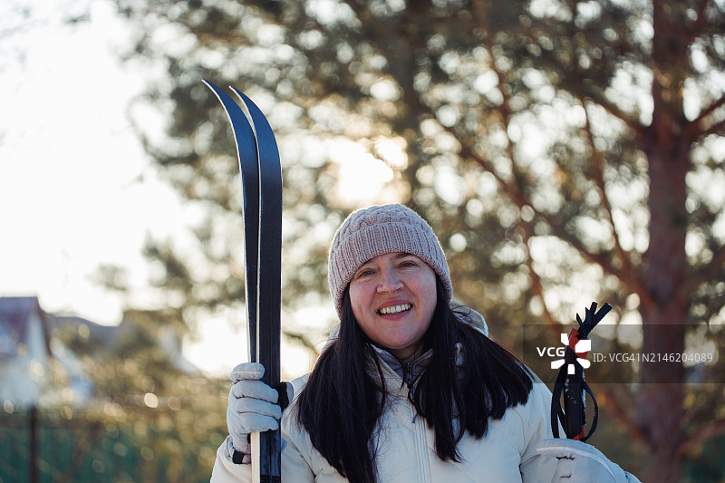 在森林里滑雪前，微笑的成熟女子拿着滑雪板和滑雪棒。图片素材