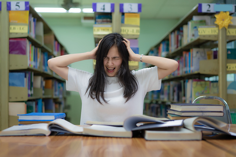 亞洲女學生無聊地在圖書館看書，大學里有很多書。亞洲學生沮喪地看書準備考試圖片下載