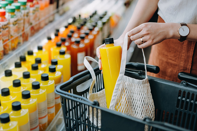 裁剪的鏡頭，年輕的亞洲婦女購物新鮮果汁冷藏貨架在超市和把一瓶新鮮榨橙汁棉花網生態袋在購物車?！傲憷速M”的概念圖片素材