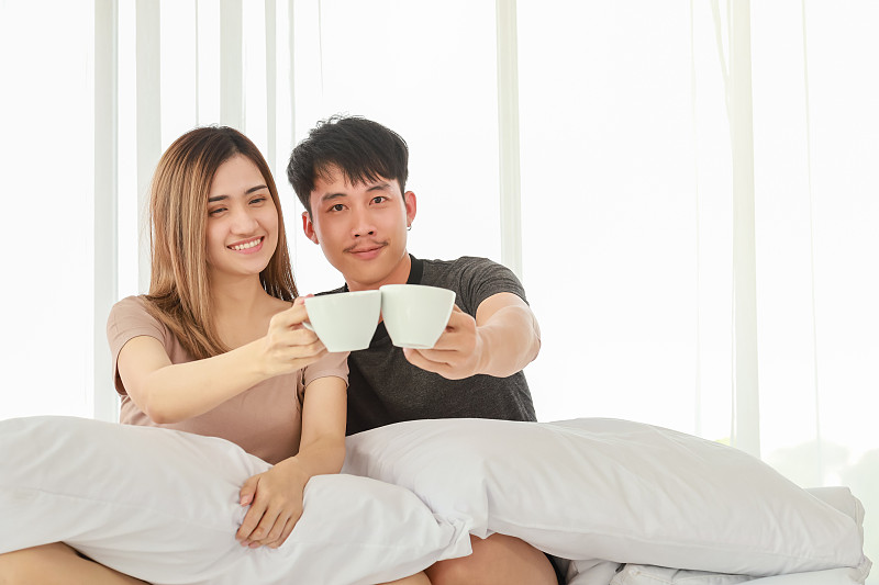 情侶節和情人節概念。亞洲男性和女性的肖像拿著白色的熱咖啡杯和坐在床上的臥室圖片下載