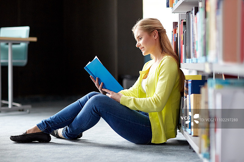 教育，高中，大学，学习和人的概念-微笑的学生女孩坐在图书馆的地板上看书图片素材