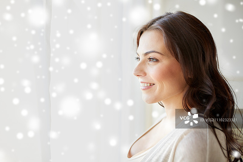 人们，冬天，圣诞节和休闲概念-快乐的年轻女子透过窗户在家里看雪图片素材