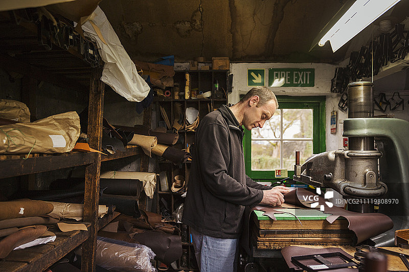 一个男人站在鞋匠的车间里，切割着棕色的皮革。图片素材