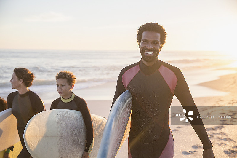 肖像微笑，自信的男性冲浪者携带冲浪板与家人在阳光明媚的夏天日落海滩图片素材