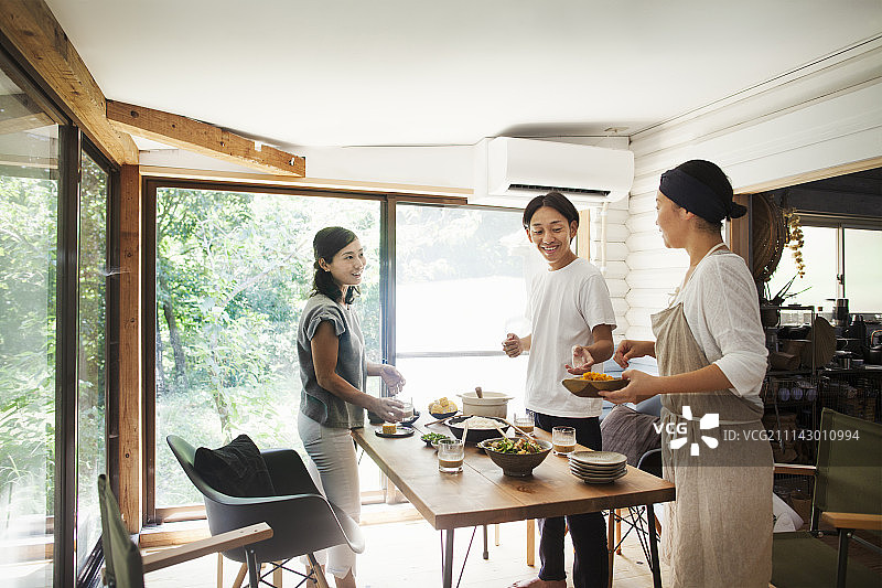 一名男子和两名微笑的女子站在摆着盘子和碗食物的桌子周围。图片素材