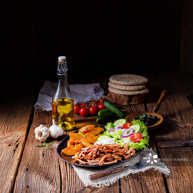 烤肉搭配薯条和新鲜的沙拉，放在黑色的盘子里，棕色的木桌上，配以新鲜的蔬菜、皮塔饼和橄榄油图片素材
