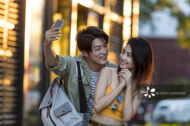 幸福情侣背着包在户外步行街旅行度假拿着手机拍照嬉戏图片素材