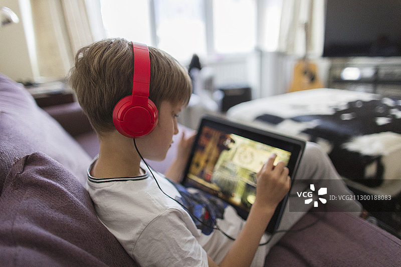 男孩戴着耳机和平板电脑在沙发上玩电子游戏图片素材