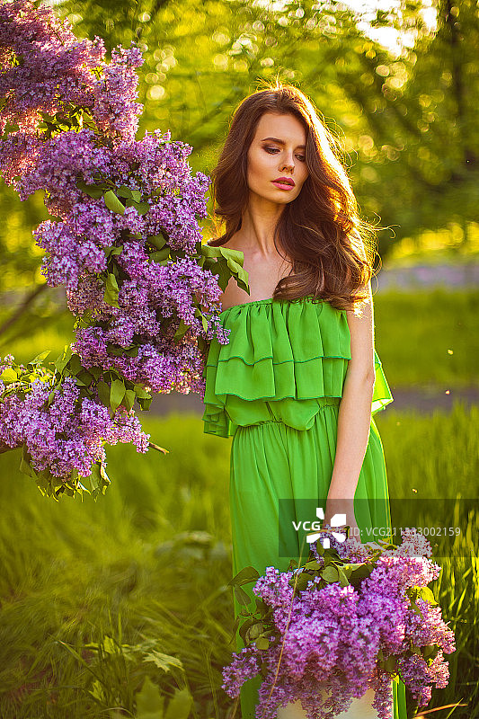 迷人的女人在绿色连衣裙摆姿势与丁香花在田野图片素材