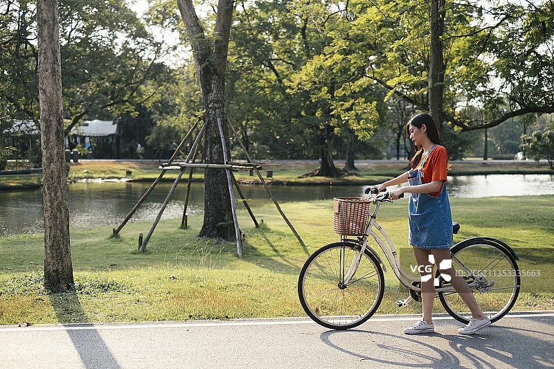 长发女孩穿着橙色t恤和牛仔裤套头衫，骑着自行车沿着公园的草地走。图片素材