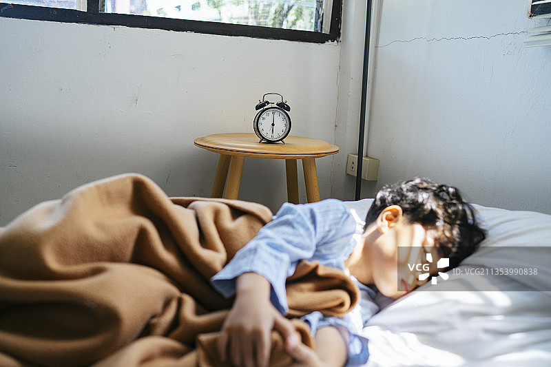 中午背着睡觉的男孩的床旁边的木桌上放着一个古董闹钟。图片素材