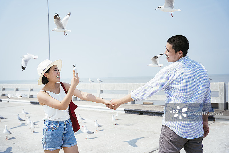 幸福浪漫的亚洲泰国情侣手牵着手，在一群海鸥中拿着智能手机自拍。图片素材