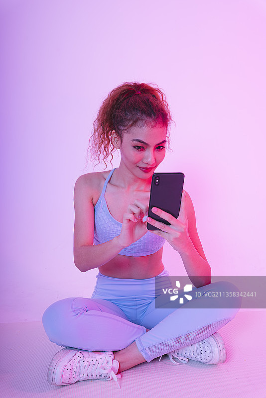 健康的年轻亚洲卷发女人的肖像在健身房使用智能手机。图片素材