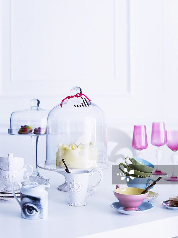 粉彩和古董风格的白瓷，蛋糕架玻璃盖和粉红色的开胃酒杯和彩色杯子在白色的桌子上图片素材