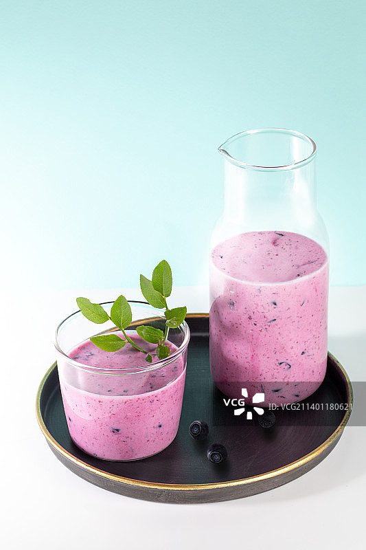 玻璃杯和一瓶蓝莓冰沙与新鲜的叶子在绿松石背景的圆形盘子，俄罗斯图片素材