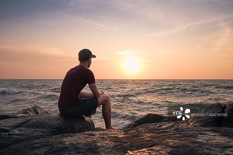 日落时，男人坐在沙滩上的岩石上，背对着天空图片素材