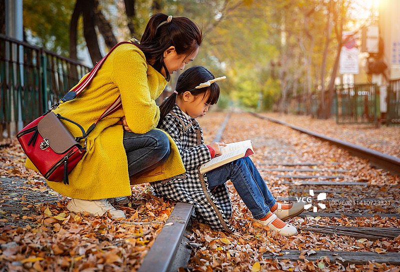 秋天户外亚洲母女坐在落满树叶的轨道上看书图片素材