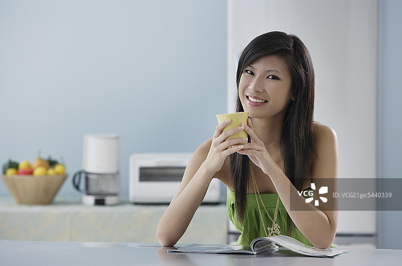 女人坐在厨房里拿着杂志和马克杯，对着镜头微笑图片素材