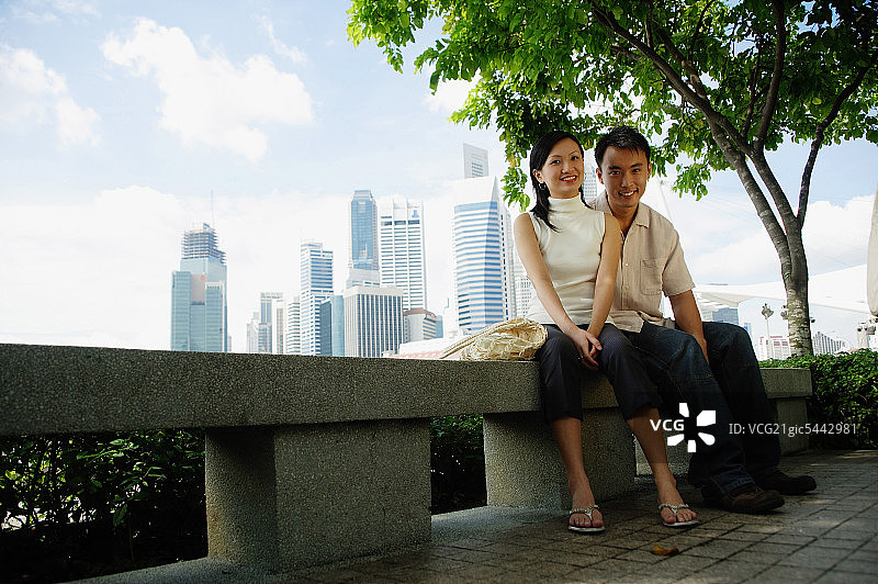 一对年轻夫妇坐在公园的长椅上，看着镜头，背景是建筑物图片素材