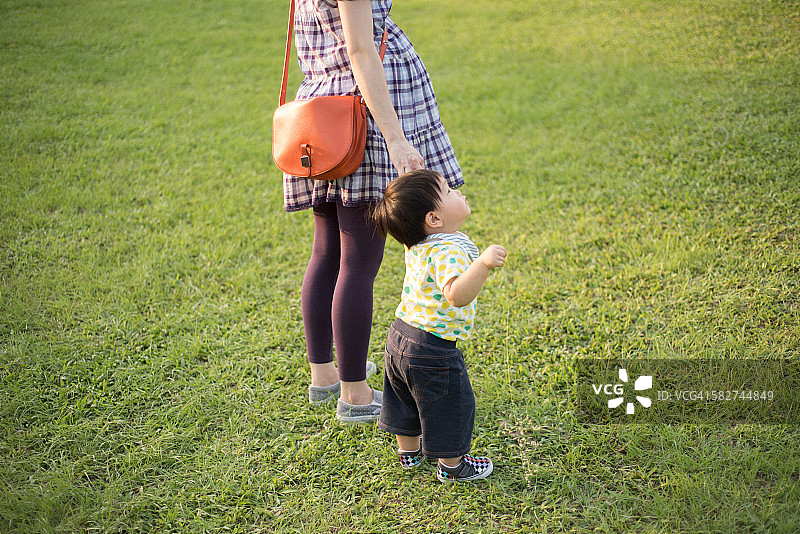 蹒跚学步的小男孩牵着妈妈的手在草坪上图片素材