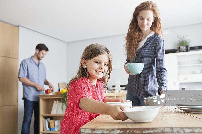 小女孩微笑着在厨房摆桌子，她的父母在后面图片素材