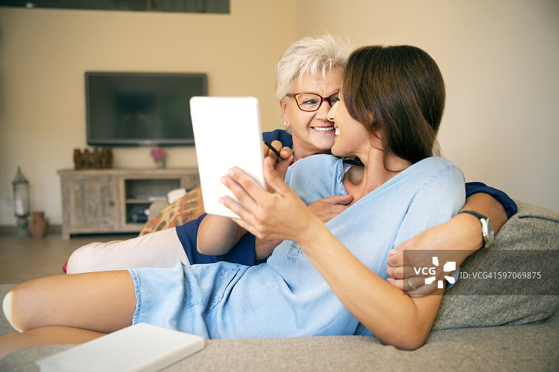 快乐的老年妇女和成年的女儿在沙发上用数码平板电脑图片素材