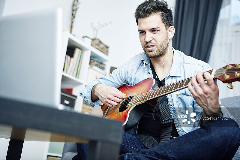 一个年轻人坐在家里的沙发上一边弹吉他一边看笔记本电脑图片素材