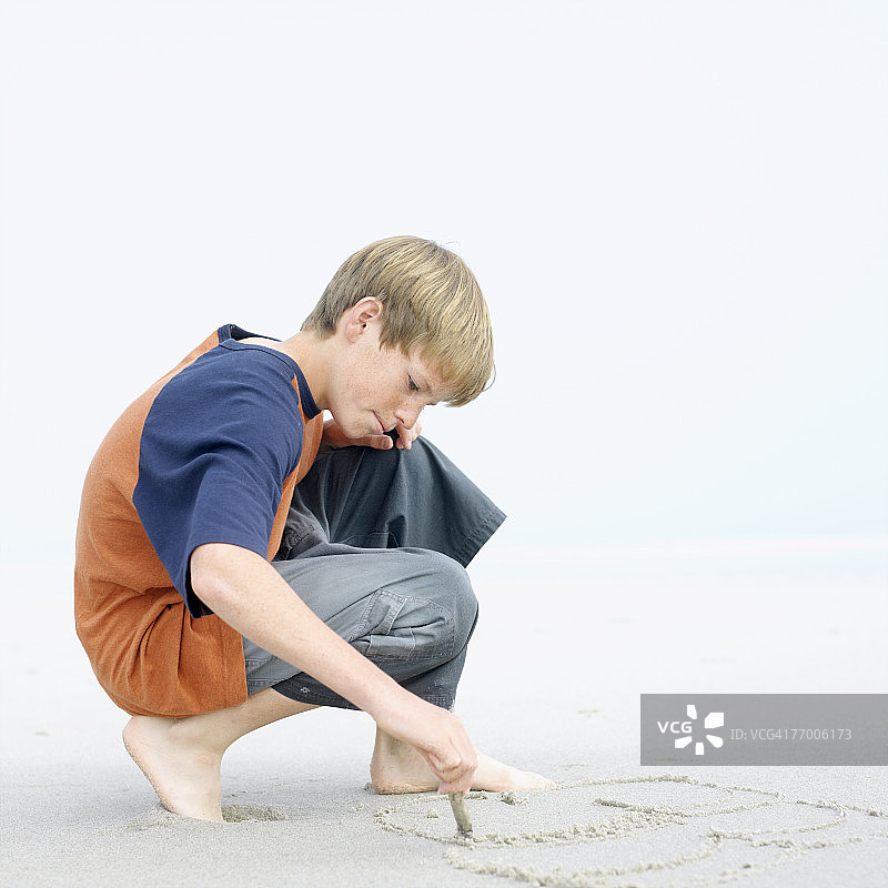 男孩(12-13岁)蹲在沙滩上，在沙滩上画画图片素材