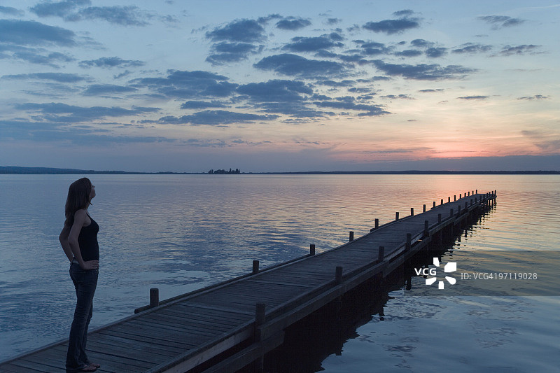 年轻女子在湖边的码头上观看雄伟的日落云景图片素材