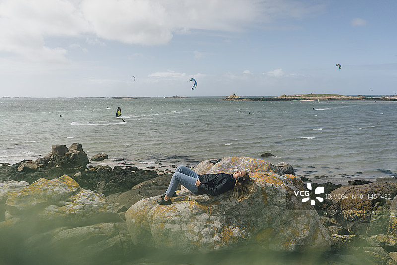 法国，布列塔尼，兰达达，年轻女子戴着耳机躺在海边的岩石上图片素材