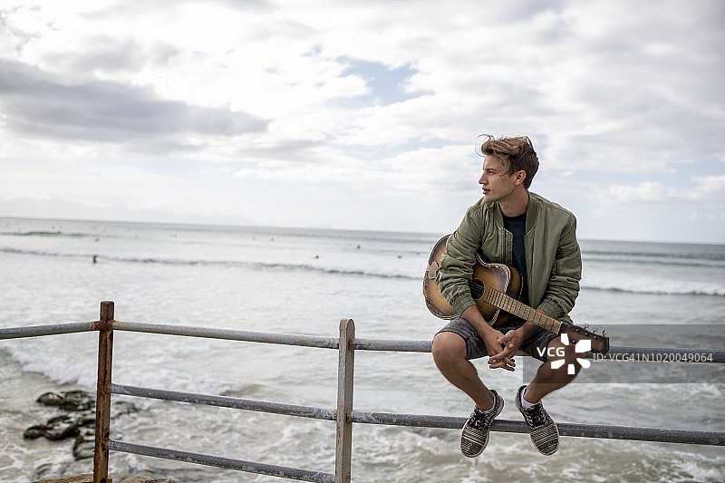 一个年轻人拿着吉他坐在海边的栏杆上看风景图片素材