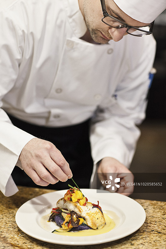在一间商业厨房里，一名白种男厨师正在对一盘鱼进行最后的润色。图片素材