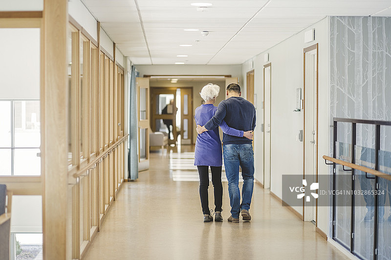 后视图的老年妇女与儿子走在走廊在疗养院图片素材