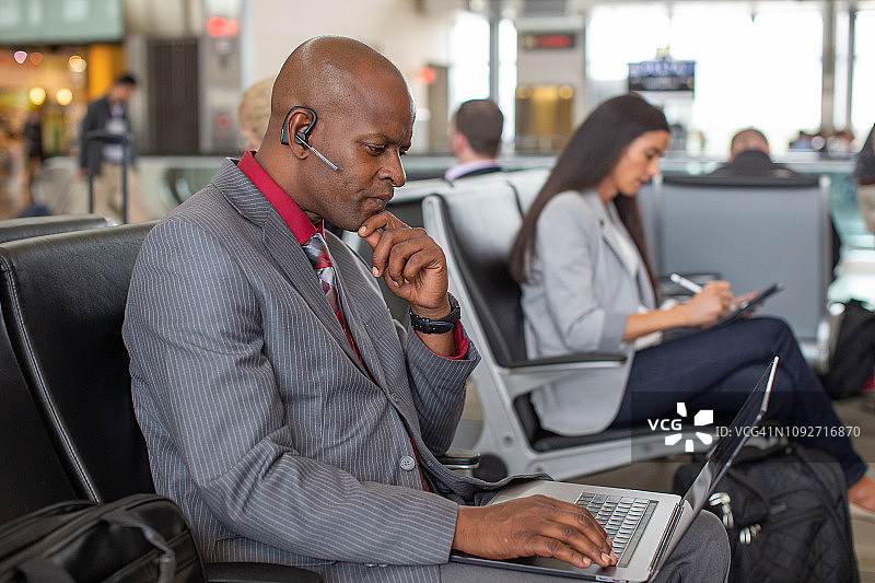 一位戴着耳机的非裔美国商人在机场候机楼门口用笔记本电脑工作。图片素材