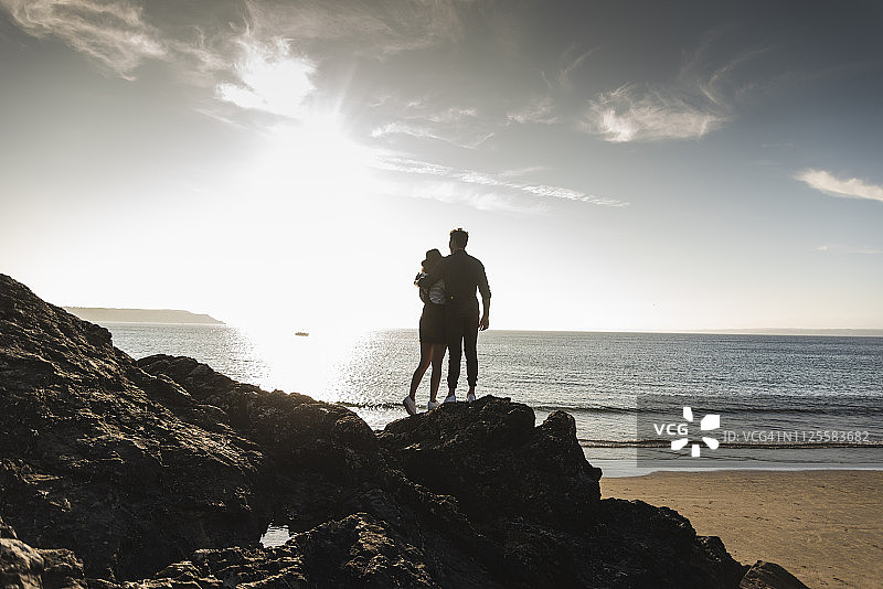 法国，布列塔尼，年轻夫妇在日落时站在海滩岩石上的后视图图片素材
