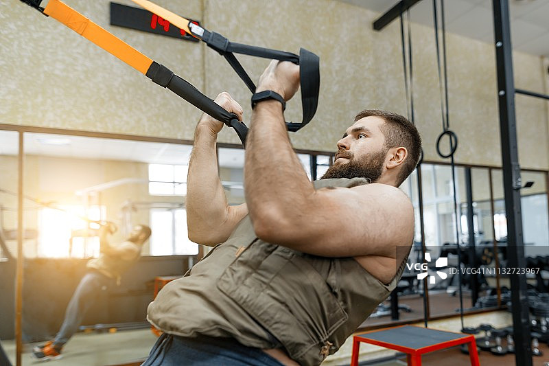 肌肉发达的胡子男子穿着军事加重装甲背心做练习使用带子系统在健身房。运动，训练，健美和健康的生活方式的概念。图片素材