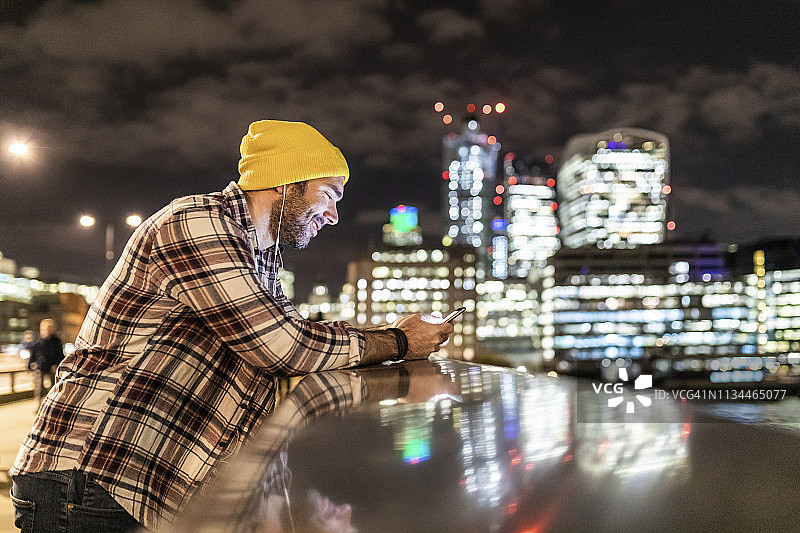 英国，伦敦，一名微笑的男子倚靠在栏杆上，看着他的手机，背景是城市的灯光图片素材