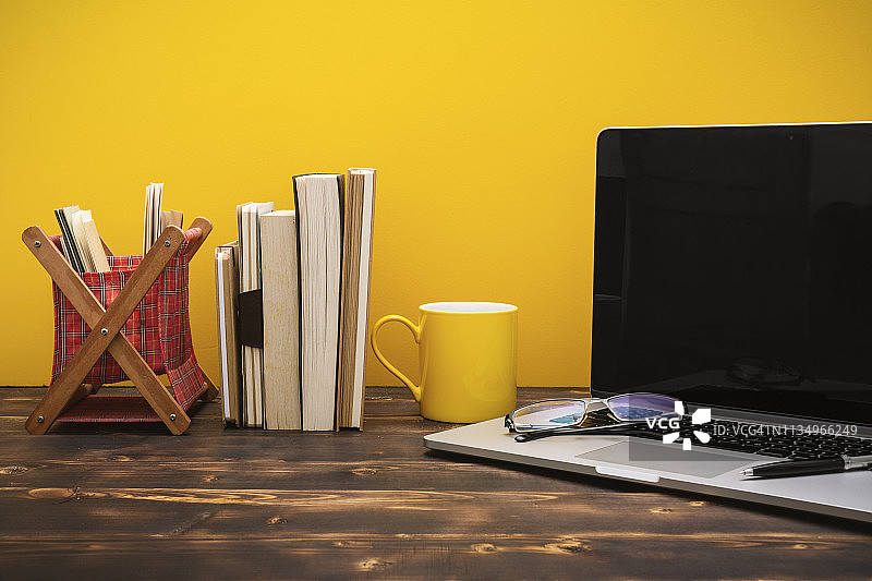 黄色背景下的木桌上，笔记本电脑和书籍排成一排图片素材