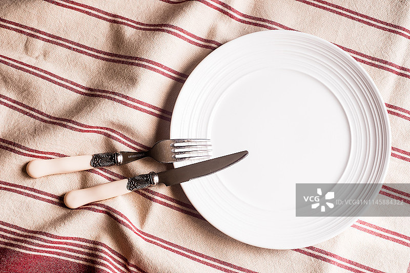 空空的白色盘子，刀叉放在红白相间的亚麻条纹餐巾上，俯视图。图像与复制空间。厨房桌子与毛巾和一个盘顶视图与复印空间。图片素材