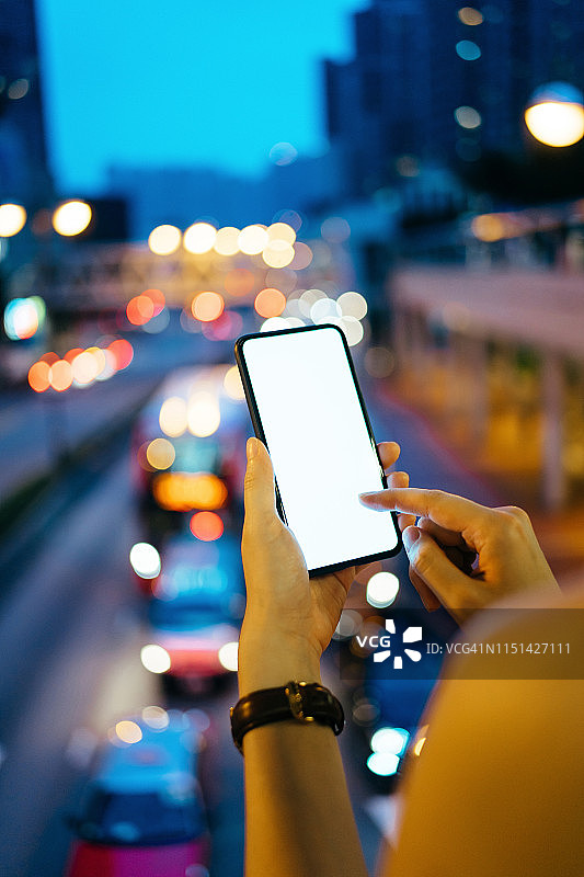 近距离的女人的手使用智能手机在户外的城市天际线与繁忙的城市交通在闹市区图片素材
