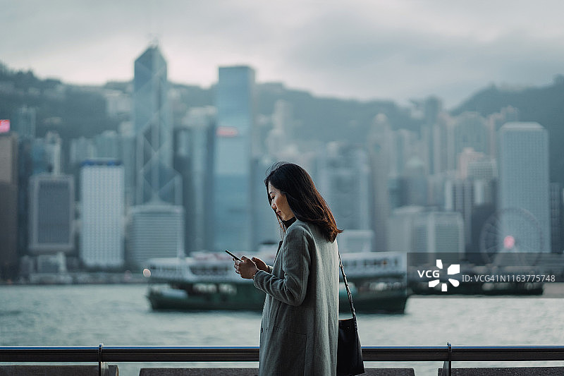 年轻的亚洲女商人在维多利亚港的海滨长廊上使用智能手机，背靠香港中央商务区的城市景象图片素材