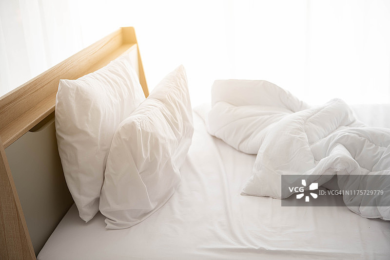白色未整理的床在早晨与阳光与白色窗帘背景在优雅的家。室内设计及豪华家具概念。室内生活方式和懒人活动主题。图片素材