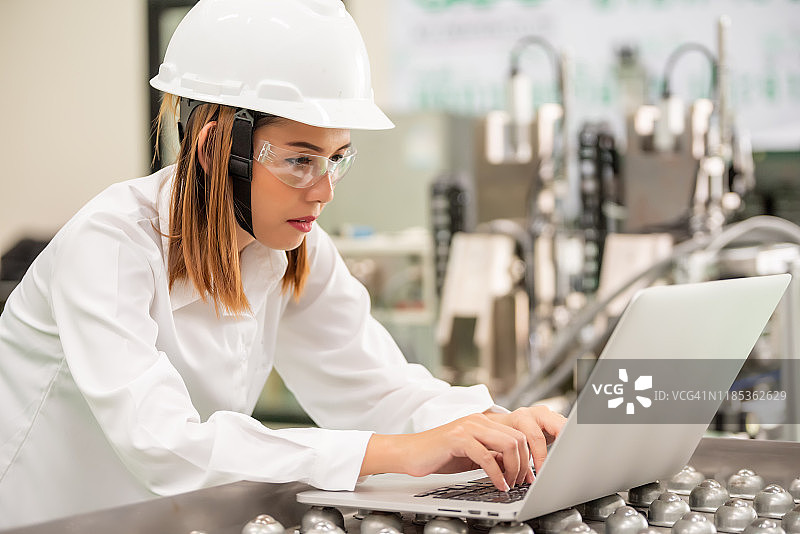 亚洲女工程师戴着安全帽和白色安全帽。同时在工厂用笔记本电脑控制机器图片素材