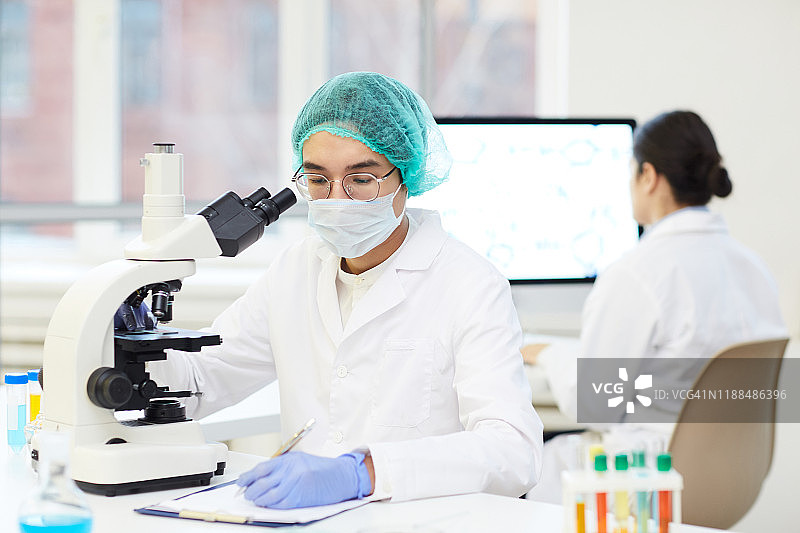 严肃年轻的亚洲实验室研究员，穿着无菌工作服，坐在书桌前，用显微镜记录样品图片素材