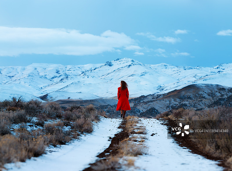 背影，年轻女子在长长的红色大衣走在雪覆盖的双轨土路，向雪山覆盖图片素材