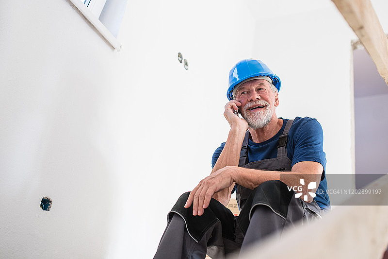 一位戴着头盔的老人坐在房屋建筑工地上，使用智能手机。图片素材