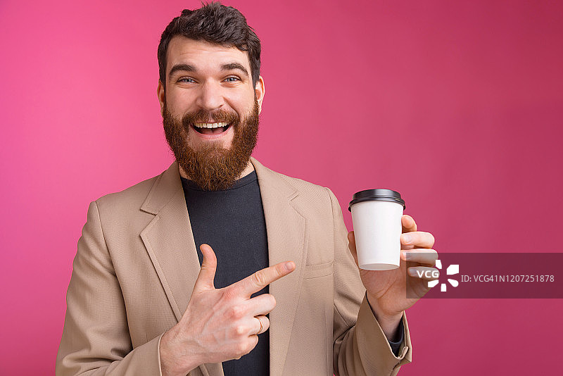 英俊的年轻大胡子男子正指着一个白色的咖啡杯去粉红色的背景。图片素材