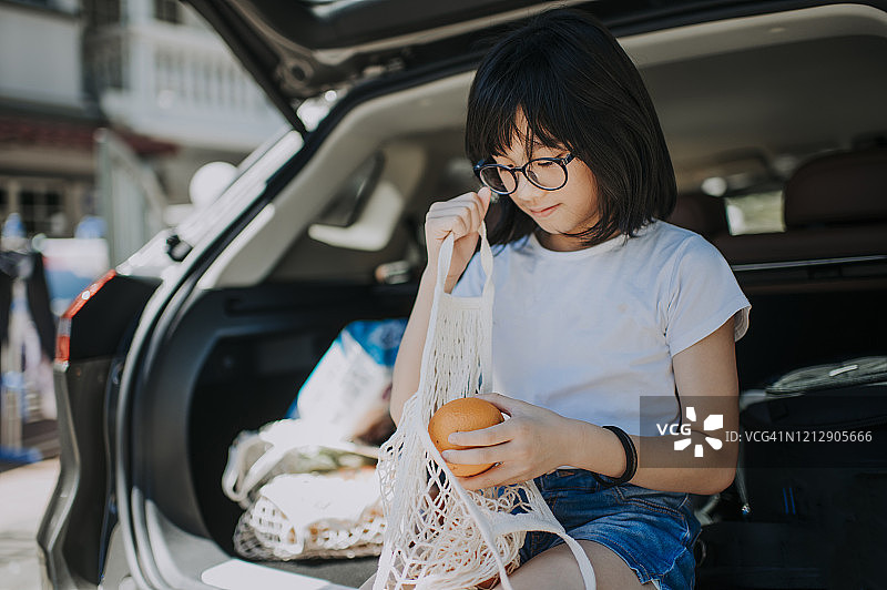 一个亚洲华裔年轻女孩坐在汽车的后备箱里，手里拿着一个橘子图片素材