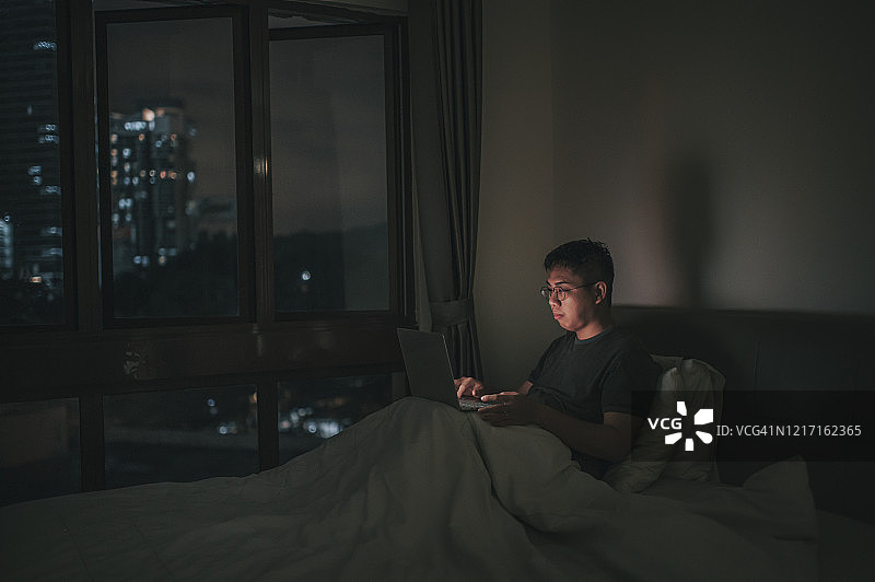 一个亚裔中国中年成年人晚上在家里的床上用他的笔记本电脑工作图片素材