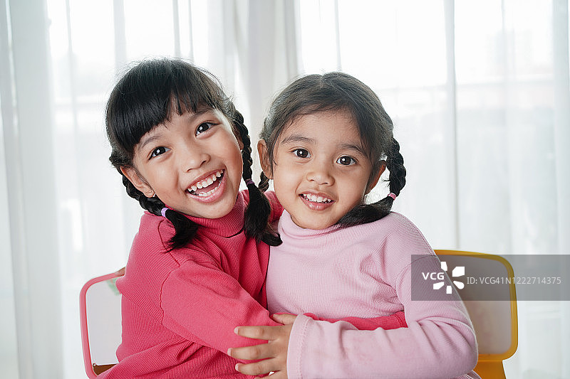 快乐的亚洲小女孩拥抱和她的快乐的妹妹或朋友在客厅在家里的乐趣。家庭和童年观念。图片素材